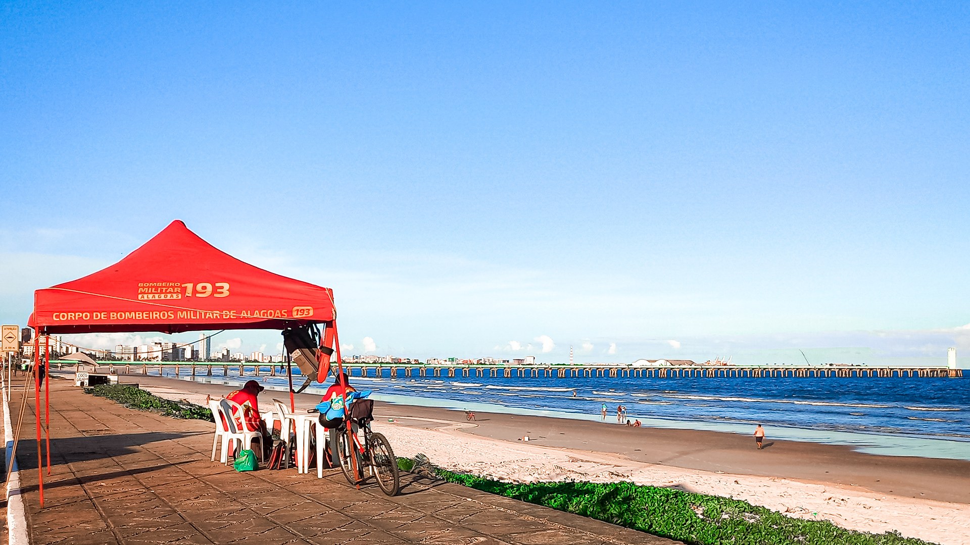 Guarita vermelha do Corpo de Bombeiros Militar de Alagoas montada no calçadão da praia de Maceió, com um bombeiro de serviço e o mar ao fundo